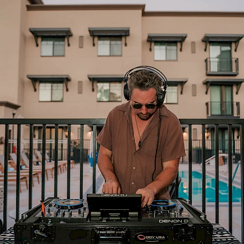 A DJ wearing sunglasses and a brown shirt spins records at a poolside setup, with a patterned table, gear, and a hotel-style building in the background.