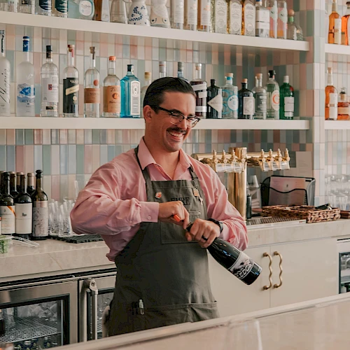 A smiling bartender in a pink shirt and apron pops a wine cork behind a well-stocked bar with shelves of bottles; cheerful, lively scene.