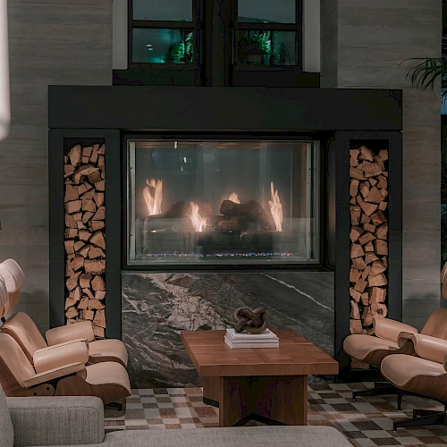 A modern living room with a lit stone fireplace centered between built-in log racks, flanked by beige leather chairs and a wooden coffee table, cozy mood.