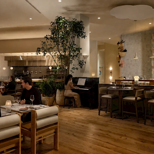 People dining in a modern, warmly lit restaurant with wooden floors, plants, and a bar area in the background.