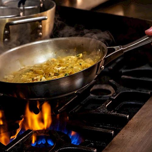 The image shows a person cooking in a frying pan over a gas stove, with flames under the pan and steam rising from the sautéing ingredients, while a hand holds the pan handle, keeping it steady.