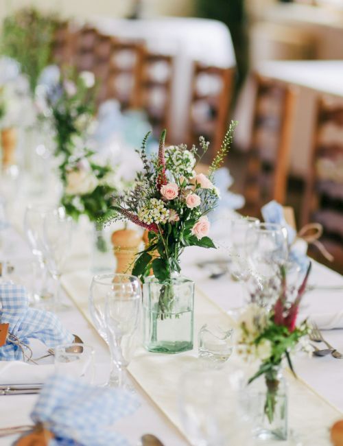 A long dining table is elegantly set with white linens, floral centerpieces, glassware, and blue checkered napkins, ready for guests.
